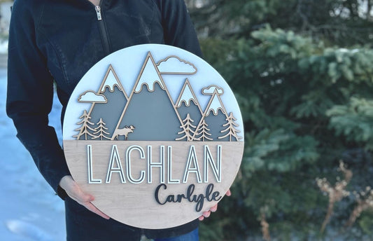a white coloured round with 4 grey mountains with snowy tops and 3 clouds around the peaks. At the base of the mountains there are trees and a moose.   The bottom half of the sign is a maple backer, sanded beautifully featuring the name "Lachlan" in a bold, yet modern print painted white. to the bottom right in a smaller cursive font it reads "Carlyle"